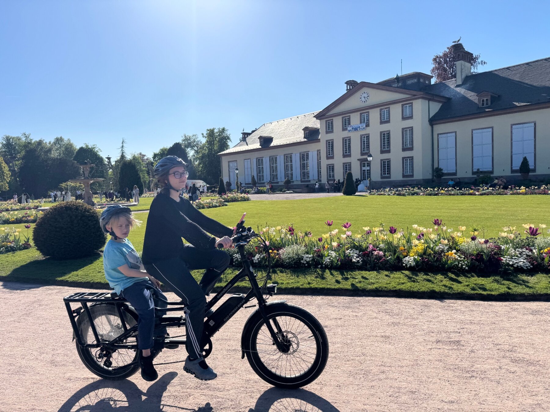 Parent and child riding a STRIM cargo e-bike past Pavillon Joséphine and tulip beds in Parc de l'Orangerie, Strasbourg
